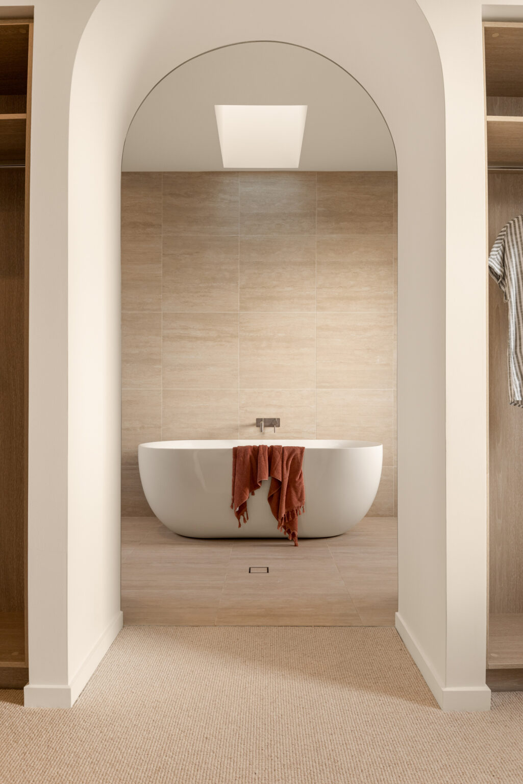 Modern bathroom with white freestanding bathtub under arched ceiling, natural wood tile walls, and neutral carpeting.