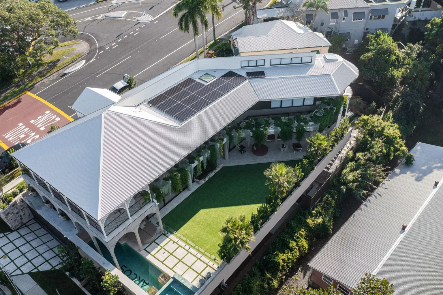 Aerial view of modern white house with solar panels, terraced garden, pool, and manicured lawn surrounded by vegetation.