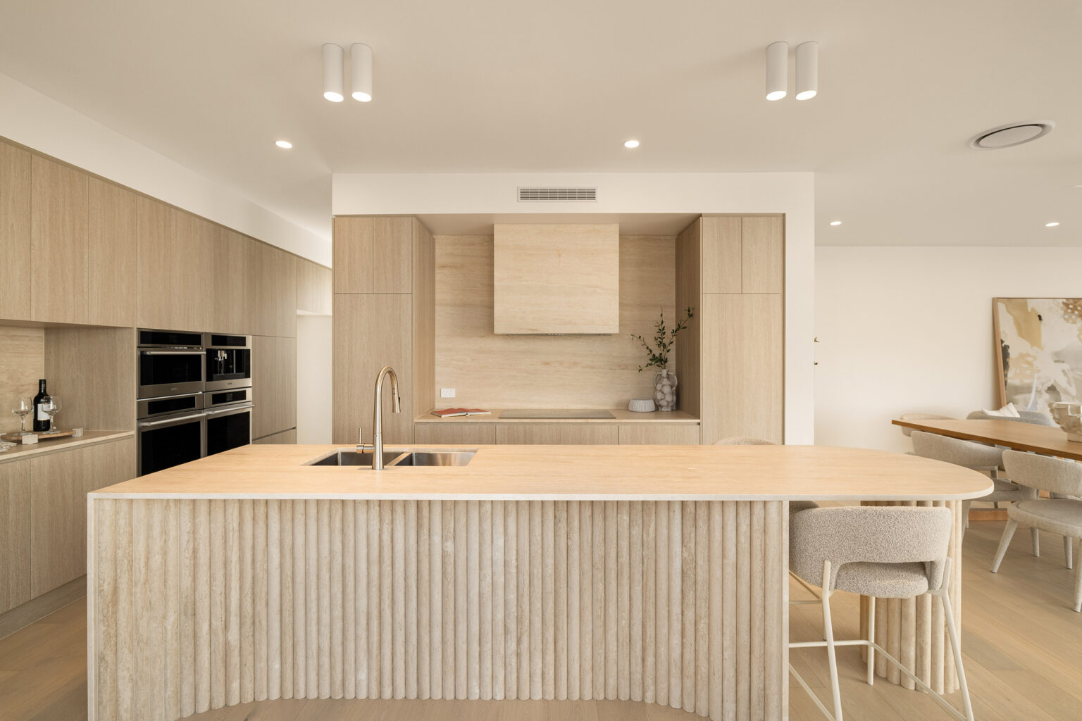 Modern kitchen with light wood cabinetry, large island with sink, built-in appliances, and minimalist design aesthetic.